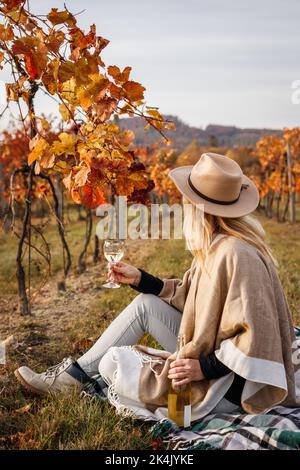 Frau mit Poncho und Hut sitzt auf der Decke und trinkt Weißwein. Entspannung im Herbst Weinberg Stockfoto