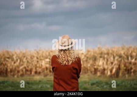 Frau mit rotem Mantel und Cowboyhut blickt auf den wolkigen Himmel über dem Maisfeld. Bäuerin auf dem Land Stockfoto