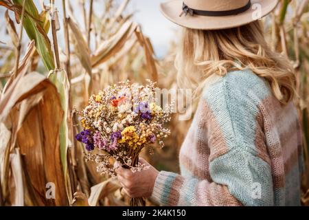 Frau mit Hut und Strickpullover mit Blumenstrauß im Maisfeld. Weibliche Mode-Kleidung für den Herbst Stockfoto
