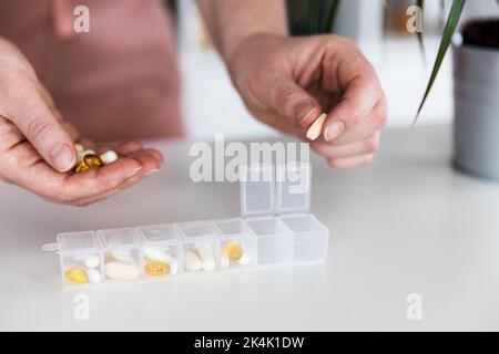 Weibliche Hände, die Pillen sortieren. Junge Frau bekommt ihre täglichen Vitamine zu Hause. Nahaufnahme der medizinischen Pillenbox mit Dosen von Tabletten für den täglichen Gebrauch nehmen Sie ein Medikament mit verschiedenen weißen, gelben Medikamenten und Kapseln Stockfoto