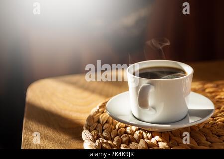 Kaffeetasse mit heißem Dampf auf braunem Holztisch mit morgendlicher Sonneneinstrahlung durch das Fenster. Gesunder schwarzer Kaffee in einem Becher. Der Kopierbereich befindet sich auf der linken Seite. Licht- und Schattenkonzept. Stockfoto