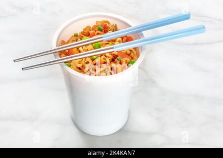 Ramen-Tasse, Instant-Soba-Nudeln in einem Plastikbecher mit Gemüse, grünen Erbsen und Karotten, mit Essstäbchen auf weißem Marmorboden Stockfoto