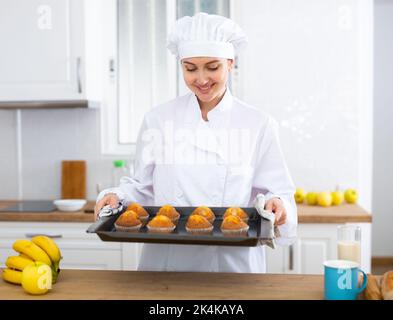 Glückliche weibliche Köchin in weißer Uniform, die mit einem Tablett mit Muffins in der eigenen Küche stand Stockfoto