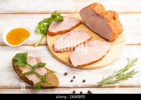 Geräucherter Schweineschinken auf Schneidebrett mit Pfeffer und Kräutern auf weißem Holzhintergrund. Seitenansicht, Nahaufnahme. Stockfoto