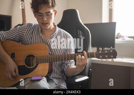 Männlicher Teenager singt Lied spielt akustische Gitarre und liest Text auf dem Smartphone-Anwendungsbildschirm Stockfoto