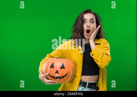 Erschrocken aufgeregt schockierte kaukasische Frau in legerer Kleidung, hält gemalten halloween-Kürbis, steht auf isoliertem grünen Hintergrund, Blick auf die Kamera in Staunen. Halloween-Party, Albtraum-Konzept Stockfoto