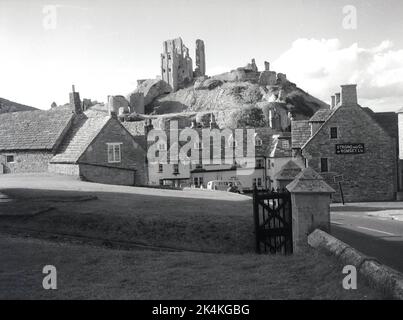 1964, historisch, das Dorf Corfe Caste und ein Blick auf das Greyhound Hotel, mit den Ruinen von Corfe Castle auf einem Hügel, Dorset, England, Großbritannien. Zeichen für Strong und Co von Romsey Ltd an der Wand des Gebäudes. Später wurde es ein Whitebrot Pub. Das Pub aus dem Jahr 1580 ist eines der ältesten Gasthöfe Englands. Ursprünglich von Wilhelm dem Eroberer erbaut, war die Burg auf dem Hügel eine der frühesten Befestigungen, die teilweise aus Stein gebaut wurden, aber 1645 wurde sie zertrümmert, d.h. nach dem englischen Bürgerkrieg teilweise zerstört, um zu verhindern, dass sie als mögliche Festung genutzt wurde und zu einer Ruine wurde. Stockfoto