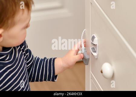 Das Baby öffnet das Kinderschloss in der geschlossenen Schublade des Schranks. Kleinkind Baby mit Hand öffnet Türschloss auf der Brust der Schubladen. Kind im Alter von einem Jahr und drei Stockfoto