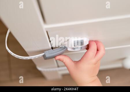 Das Baby öffnet das Kinderschloss in der geschlossenen Schublade des Schranks. Kleinkind Baby mit Hand öffnet Türschloss auf der Brust der Schubladen. Kind im Alter von einem Jahr und drei Stockfoto