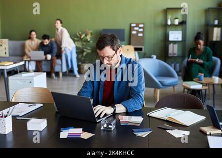 Junger seriöser männlicher Designer, der beim Ansehen von Online-Videos oder Schulungen zum Thema seines neuen Projekts auf den Laptop-Bildschirm schaut Stockfoto