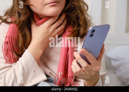 Eine kranke Frau, in einen roten Schal gewickelt, mit einem Telefon in der Hand, Kehlschmerzen. Erwachsene kranke Frau, die auf einem Heimbett in einem weißen Schlafzimmer sitzt, Frau im Alter von 35 Jahren Stockfoto