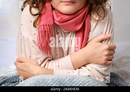 Eine Frau friert von der Kälte, die auf einem Heimbett sitzt, Fieber und hohen Temperaturen. Erwachsene kranke Frau in einem roten Schal und Decke in einem weißen Bett gewickelt Stockfoto