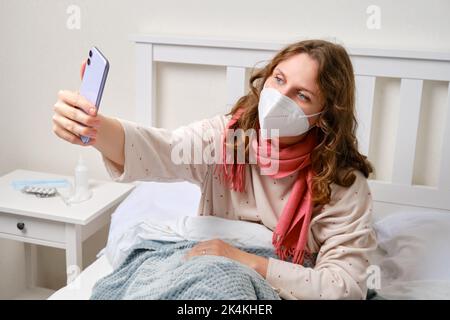 Kranke Bloggerin nimmt Selfie-Foto am Telefon für Online-Blog. Erwachsene kranke Frau in einem roten Schal gewickelt sitzt auf einem Heimbett in einem weißen Schlafzimmer, Frau A Stockfoto