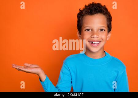 Foto-Porträt von niedlichen kleinen Schuljunge Palm halten leeren Raum toothy Lächeln tragen trendige blaue Kleidung isoliert auf orangefarbenem Hintergrund Stockfoto