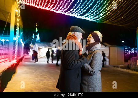 Glückliches älteres Paar, das die Hände hält und sich unter den Weihnachtslichtern wohlfühlt Stockfoto