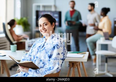 Glückliche junge Geschäftsfrau mit offenem Buch sitzt am Arbeitsplatz gegen eine Gruppe von Kollegen diskutieren Arbeitspunkte Stockfoto