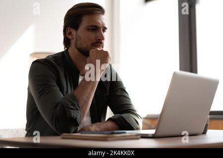 Nachdenklicher bärtiger Mann, der am Tisch sitzt, denkt am Laptop an die Problemlösung, nachdenklicher männlicher Mitarbeiter, der über die Idee nachdenkt, den Computer zu betrachten Stockfoto