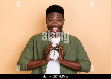 Foto von beeindruckt Millennial Brunet Kerl aufwachen in der Zeit tragen khaki Shirt isoliert auf beige Farbe Hintergrund Stockfoto