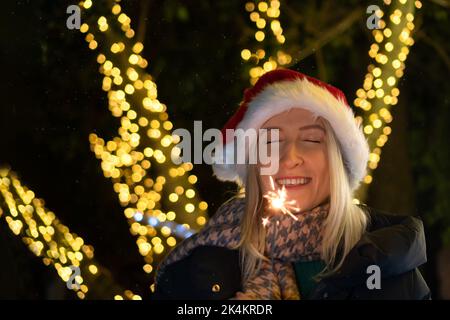 Fröhliche lächelnde Frau in einem Weihnachtsmannhut mit geschlossenen Augen hält Wunderkerzen fest und wünscht sich auf der Straße vor dem Hintergrund der nächtlichen Lichter Stockfoto