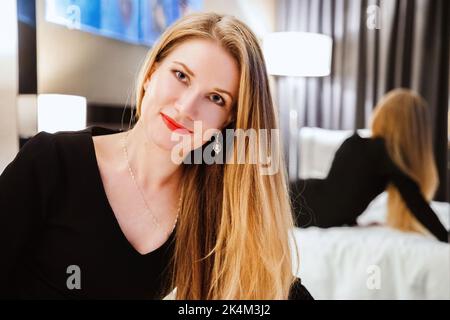 Hübsche Hündin mit langen blonden Haaren, in einem schwarzen Kleid, sitzend auf dem Bett, schaut uns an. Ihre Reflexion ist im Spiegel hinter ihr sichtbar Stockfoto