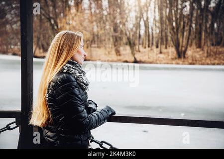 Das hübsche blonde Mädchen, das einen schwarzen Mantel, Handschuhe und Schal trägt, blickt über einen gefrorenen See; Bäume im Hintergrund Stockfoto