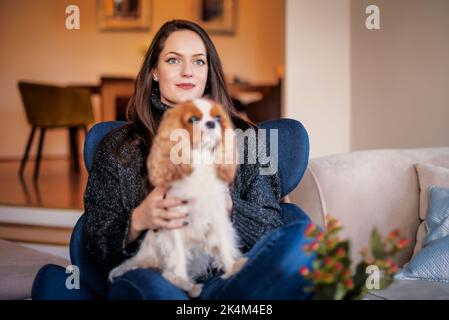 Attraktive braune haarige junge Frau, die sich zu Hause mit ihrem niedlichen Welpen entspannt. Stockfoto