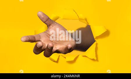 afroamerikanischer Mann, der die Handfläche in zerrissenem gelbem Papier durch ein Loch ausstreckt, helfende Hand anbietet und um Wohltätigkeit bittet Stockfoto