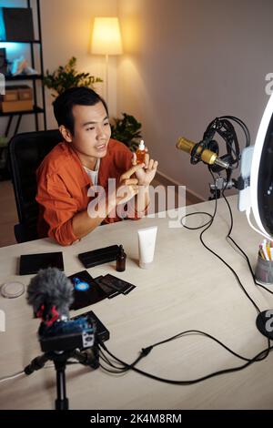 Vertikale Hochwinkelansicht eines jungen erwachsenen asiatischen männlichen Influencers, der neue Kosmetikprodukte überprüft und feuchtigkeitsspendendes Serum oder nährendes Öl zeigt Stockfoto
