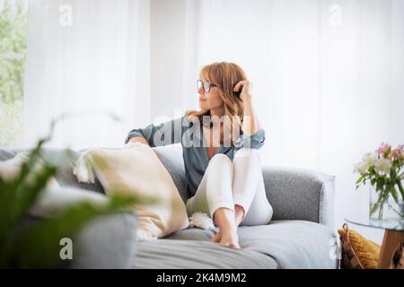 Die ganze Länge der Frau mittleren Alters entspannt sich auf der Couch zu Hause. Attraktive weibliche tragen legere Kleidung und Brillen. Nachdenkliche Frau schaut weg. Stockfoto