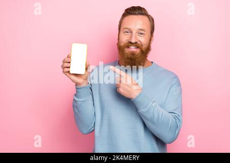 Porträt von zufrieden positive rote Haare Kerl gekleidet blauen Pullover Finger Demonstration App in Smartphone isoliert auf rosa Hintergrund Stockfoto