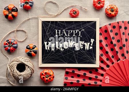 Text Frohes Halloween mit Spinnweben auf Kreidetafel. Herbstliche Einrichtung um eine Tafel. Orange Mini Kürbisse schwarz lackiert und Fächer mit Tupfen auf beige Stockfoto