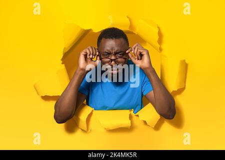 Schwarzer Mann mit schlechter Sehkraft, der eine Brille trägt und Augen schielt und durch ein Loch in gelbem Papier auf die Kamera schaut Stockfoto