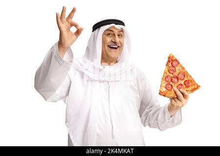 Reifer arabischer Mann in ethnischer Kleidung, der eine Scheibe Pepperoni Pizza hält und auf weißem Hintergrund ein gutes Zeichen gestikelt Stockfoto