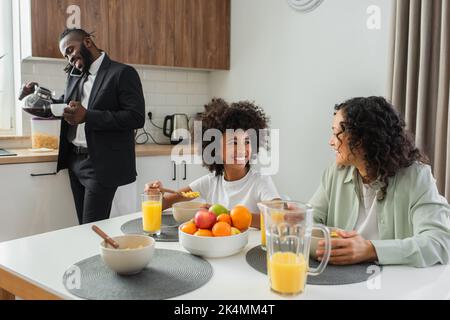 Glückliche afroamerikanische Mutter und Tochter, die einander in der Nähe von Geschäftsmann beim Frühstück auf dem Smartphone sprechen, Stockbild Stockfoto