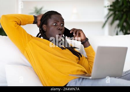 Unerwartete Neuigkeiten. Schockiert Black Guy Mit Laptop Im Gespräch Auf Handy Zu Hause Stockfoto