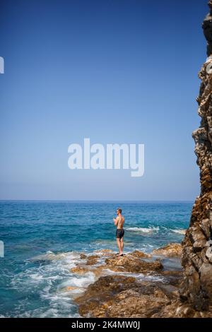 Ein junger Mann steht auf den Felsen mit Blick auf das offene Mittelmeer. Ein Kerl an einem warmen Sommer sonnigen Tag schaut auf die Meeresbrise Stockfoto