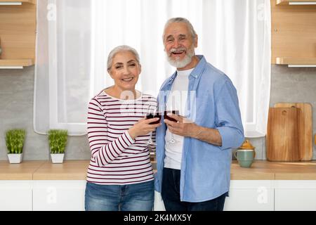 Lächelnde Ältere Ehepartner Halten Gläser Mit Wein, Während Sie In Der Küche Posieren Stockfoto