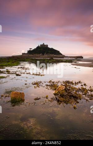 Vertikale Aufnahme von Wasser und Unkraut mit St. Michael's Mount, Cornwall und einem rosa bewölkten Himmel im Hintergrund. Stockfoto