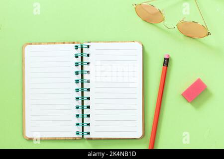 Offenes Notizbuch, leeres Papier, Bleistift, Radierer, Sonnenbrille auf pastellgrünem Hintergrund. Leerer spiralförmiger Notizblock, Kopierbereich, Draufsicht Stockfoto