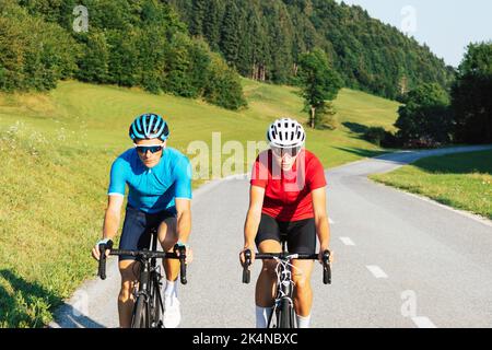 Der professionelle Rennradfahrer paart sich in Sporttrikots, trägt Fahrradhelme und Sonnenbrillen, die auf einer asphaltierten Straße treten Stockfoto