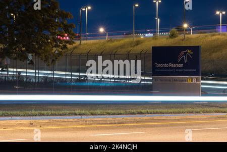 Ein Hinweisschild zum internationalen Flughafen Toronto Pearson. Flughafenterminals, die bei Nacht mit Lichtstrecken in der Umgebung zu sehen sind. Stockfoto