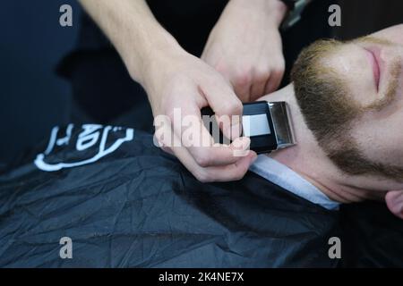 Der professionelle Meisterbarbier rasiert den Bart des Kunden mit einem elektrischen Trimmer. Haarschnitt des Bartes eines Mannes in einem Friseurladen. Barbier Men. Werbung und b Stockfoto