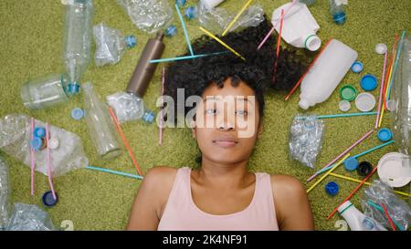 Afroamerikanische Frau liegt auf flauschigem grünen Teppich in der Nähe von verstreutem Plastikmüll. Junge Hausfrau auf dem Boden sieht in Kamera obere Nahansicht Stockfoto