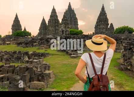 Frau, die während des Sonnenuntergangs auf ein Wahrzeichen am Prambanan Tempel schaut. Asiatische Frau mit weißem Kleid und Hut. Stockfoto