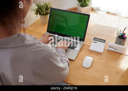 Junge Geschäftsfrau oder Freiberuflerin tippen auf dem Tastenfeld des Laptops mit leerem grünen Bildschirm, während sie die neue mobile Anwendung verwenden Stockfoto