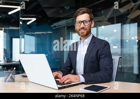 Porträt eines reifen Chefs im Büro mit Laptop, eines erfolgreichen und zufriedenen Investorenmanagers, der die Kamera anschaut, eines lächelnden Mannes in Brille und Business-Anzug, eines Investors mit Bart auf einem Stuhl Stockfoto