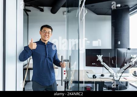 Porträt eines asiatischen Designers, eines lächelnden Mannes und eines bejahend haltenden Mannes, eines Geschäftsmanns, der mit mehreren Computern im verdrehten Bürogebäude arbeitete. Stockfoto