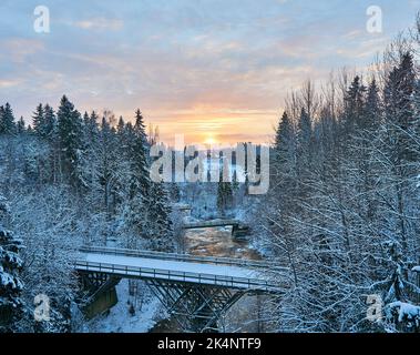 Eine Luftaufnahme einer Brücke über einen Fluss, die während des Sonnenuntergangs durch einen schneebedeckten Wald führt Stockfoto