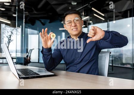 Unzufriedener asiatischer Geschäftsmann mit dem Finger, schlechte Geste des Verlusts, mit Bitcoin-Geld in virtueller Währung, Mann, der enttäuscht auf die Kamera schaut und im modernen Bürogebäude arbeitet. Stockfoto