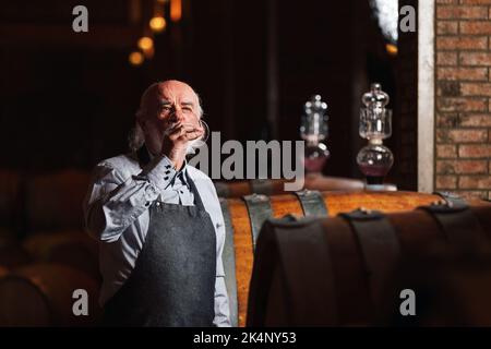 Kaukasischer Senior-Graubart-Winzer, der Rotwein verkostet, um seine Qualität und seinen Geschmack zu ermitteln, steht in der Nähe eines großen Eichenfasses Stockfoto